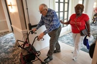 Resident Eugene Preslier, right, is accompanied by his private certified nursing aid Miriam Louis as he returns from a meal to his apartment in the independent living facility at the Toby and Leon Cooperman Sinai Residences, July 4, 2025, in Boca Raton, Fla. (AP Photo/Rebecca Blackwell)